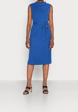 Vestido azul sin mangas con cuello tipo mock, cintura ceñida con cinturón, bajo hasta la rodilla y tela lisa. Combinado con sandalias negras de tacón grueso.