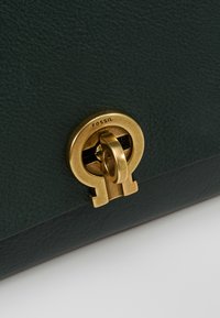 Borsa in pelle verde scuro con finitura texturizzata, dotata di una chiusura centrale in metallo dorato circolare contrassegnata "FOSSIL".