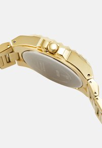 Reloj de metal dorado con un bisel texturizado, caja redonda y pulsera de eslabones. Presenta una corona texturizada y una parte posterior de la caja lisa y reflectante.