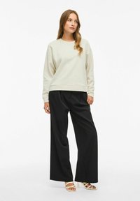 Pullover beige chiaro con la scritta "TRUTH" in rilievo, abbinato a pantaloni a gamba larga neri a vita alta e sandali bianchi con lacci.