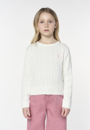 Ragazza bionda con capelli lunghi, che indossa un maglione bianco a trecce e pantaloni rosa, in piedi davanti a uno sfondo chiaro e semplice.