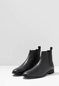 Ett par svarta läder Chelsea-boots med elastiska sidopaneler och dragögon på en vit yta mot en grå bakgrund.