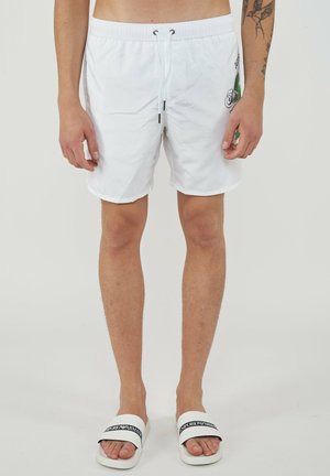 Homme portant un short blanc à cordon avec un motif latéral et des sandales blanches avec le logo noir Emporio Armani, debout devant un fond uni.