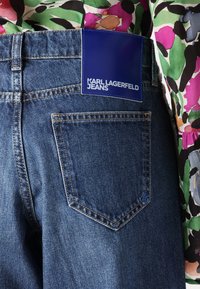 Tamsiai mėlynos džinsinės kelnes su mėlynu etiketu, ant kurio užrašyta "KARL LAGERFELD JEANS". Ypatybės: standartinės kišenės gale ir šiek tiek nudilusio audinio tekstūra.