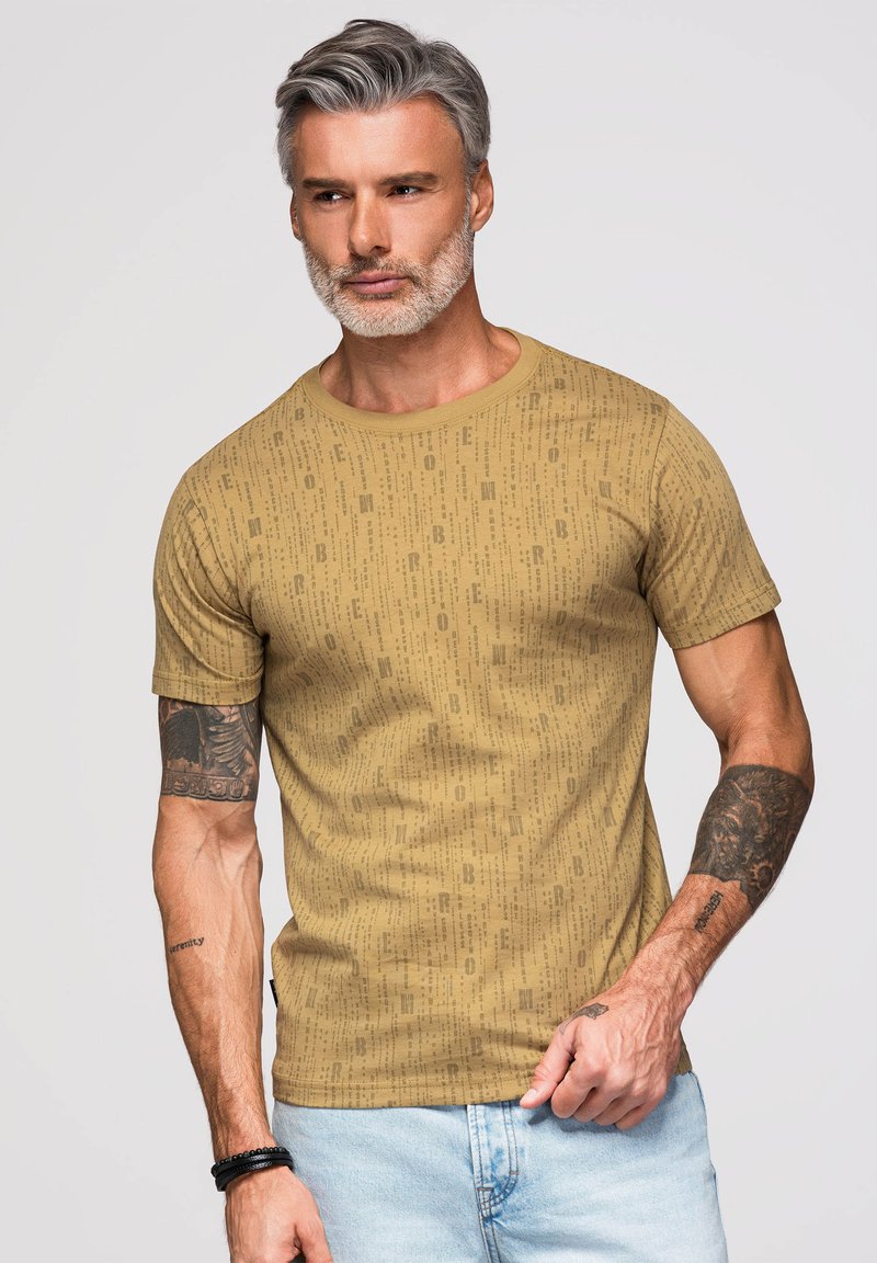 Homme aux cheveux gris et à la barbe portant un t-shirt graphique beige et un jean bleu clair, montrant des tatouages sur les deux bras avec une expression neutre.