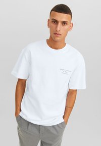 Camiseta de algodón blanca con cuello redondo y mangas cortas. Presenta un pequeño gráfico negro impreso en el lado izquierdo del pecho. Diseño minimalista.