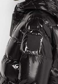 Černá péřová bunda s lesklým povrchem, s kulatou ná šivkou na rameni a žebrovanými detaily podél švů.