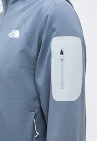 Μπλε μπουφάν North Face με υφή υφάσματος, λευκή τσέπη με φερμουάρ στο πάνω μέρος του μανικιού και λευκό λογότυπο στο στήθος.