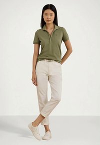 Γυναίκα με ελαιόχρωμο polo, μπεζ παντελόνι με ζώνη και ανοιχτό μπεζ αθλητικά παπούτσια, στέκεται με το ένα πόδι σταυρωμένο πάνω στο άλλο.