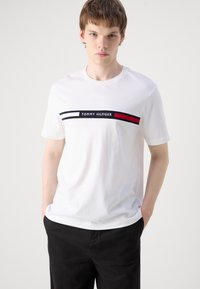 Junger Mann trägt ein weißes Tommy Hilfiger T-Shirt mit Logostreifen und schwarze Hose, steht mit den Händen in den Taschen vor einfarbigem Hintergrund.