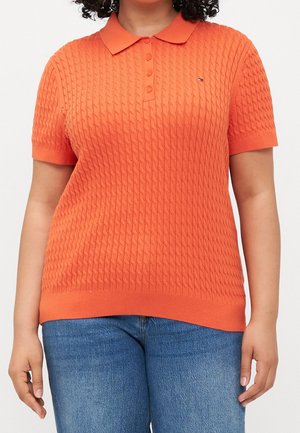 Kvinde iført en kortærmet orange polo med kabelstrik kombineret med blå jeans mod en ensfarvet baggrund.