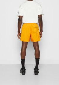 Żółte szorty z kieszenią po prawej stronie, elastycznym pasem i luźnym krojem, połączone z czarnymi skarpetkami i trampkami.