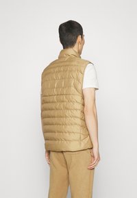 Beige quiltad pufferväst med hög krage, ärmlös design och hög hals, med en slät nylonstruktur och sidofickor.