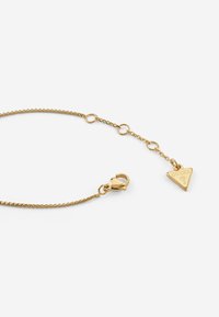 Bracciale in metallo dorato con design a catena, dotato di un ciondolo a triangolo incastonato con la scritta "GUESS" e chiusura a moschettone.