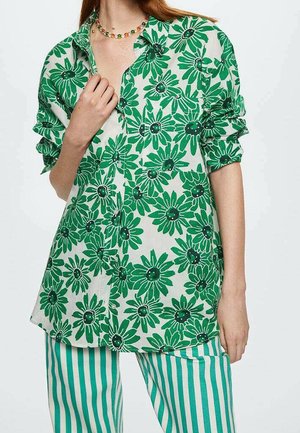 Green floral blouse with large, bold daisies on a white background. Features button closure and rolled sleeves, paired with striped green pants.