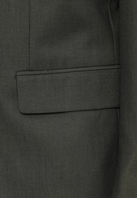 Maßgeschneiderte Blazer aus dunkelgrünem Stoff, mit einer eckigen aufgesetzten Tasche, die eine glatte Textur und ein dezentes diagonales Muster aufweist.