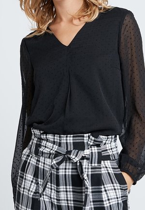 Vrouw die een zwarte doorschijnende blouse met lange mouwen met subtiele stippen draagt en een hooggetailleerde zwart-witte geruite broek, vastgebonden met een stoffen riem.
