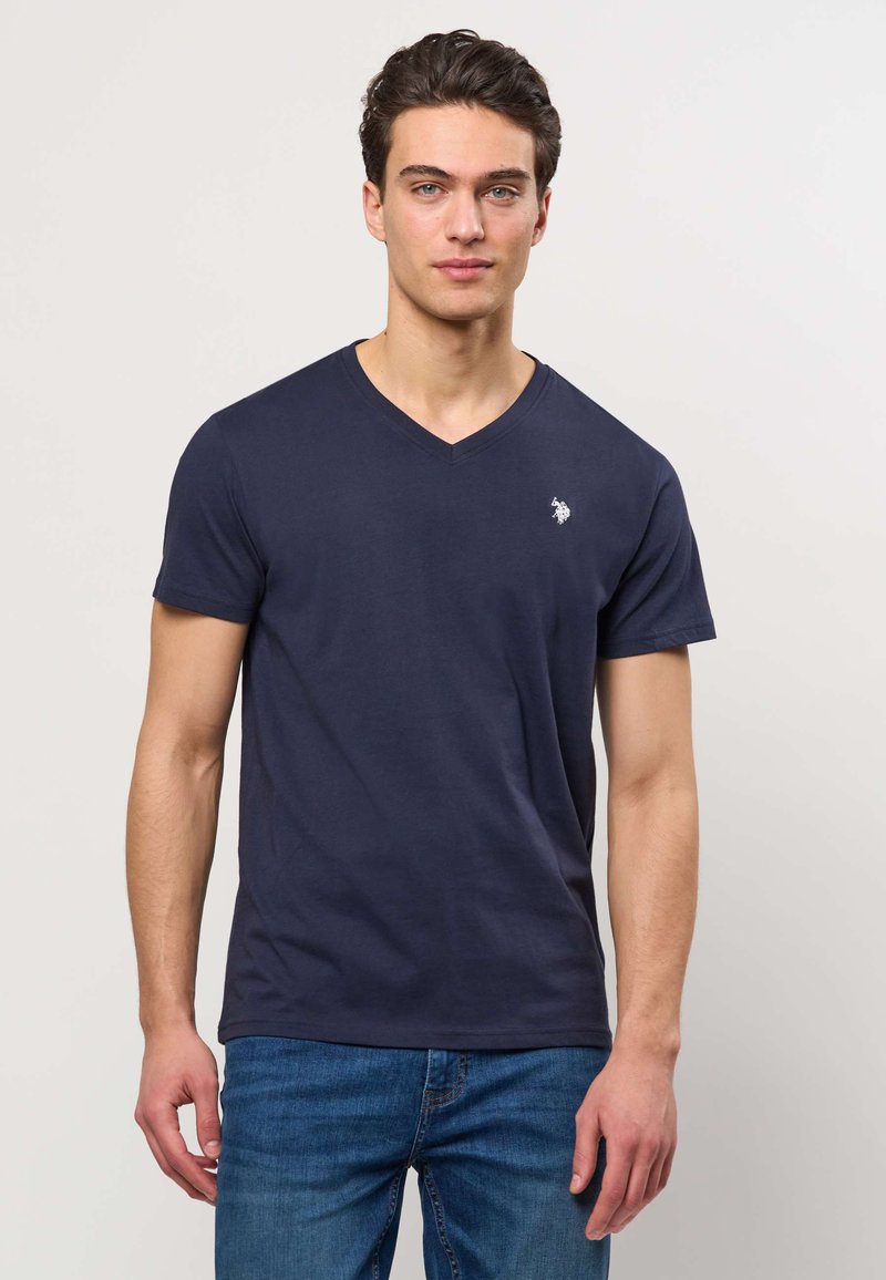 Mlad moški nosi mornarsko modro majico z V-izrezom z majhnim belim znakom na prsih in modre kavbojke, stoji pred enobarvnim svetlim ozadjem.