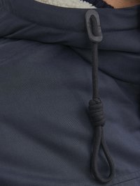 Der Kragen der dunkelblauen Jacke verfügt über einen schwarzen Kordelzug mit einem Stopper, der mit plüschigem Futter texturiert ist. Außenstoff ist sichtbar und glatt.