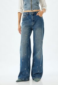 Los jeans de mezclilla azul de pierna ancha presentan detalles de ojales y una cintura alta. La tela tiene una textura clásica lavada y un diseño de cinco bolsillos.