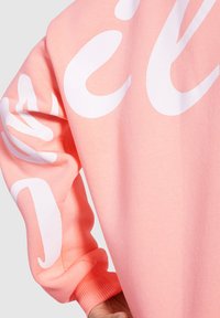 Sudadera rosa claro hecha de algodón suave, con grandes letras gráficas blancas en la manga y un detalle de puño acanalado.