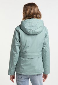 Vrouw met schouderlang golvend haar in een lichtblauwe gewatteerde jas en lichtblauwe spijkerbroek, met de rug naar de camera gericht tegen een effen achtergrond.