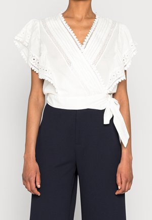 Blouse blanche enveloppante avec détails en dentelle et œillets, manches volantées, taille nouée, portée avec un pantalon noir taille haute.