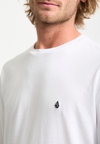Weißes Baumwoll-T-Shirt mit Rundhalsausschnitt, das ein kleines schwarzes gesticktes Logo auf der linken Brustseite aufweist, eine glatte Textur hat und einen lockeren Fit bietet.