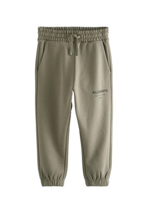 Dreng iført olivengrøn AllSaints sweatshirt og joggingsæt, hvide sneakers og sort kasket, står med hænderne i lommerne og kigger ned.