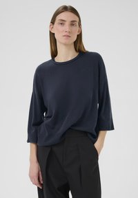 Marineblauer Strickpullover mit rundem Ausschnitt, überschnittenen Schultern und Dreiviertelärmeln. Weiche Textur und lockere Passform kombiniert mit schwarzen Hosen.
