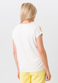 Haut blanc à manches courtes avec un col rond, texture lisse et accents à motifs sur les épaules. Associé à un pantalon jaune.