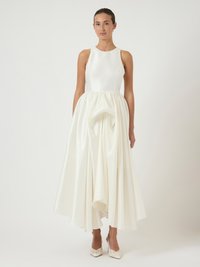 Γυναίκα που στέκεται σε μια λευκή αμάνικη τουαλέτα με εφαρμοστό κορσέ και φουσκωτό, ριχτό φούστα, φορώντας λευκά μυτερά τακούνια.