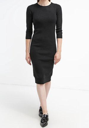 Femme portant une robe en maille noire ajustée à manches trois-quarts et des mocassins noirs brillants, debout devant un fond blanc.