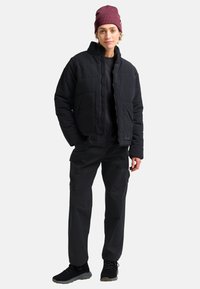 Persona che indossa una giacca a vento nera, pantaloni neri, scarpe nere e un berretto bordeaux, in piedi con le mani in tasca.