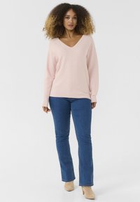 Svetlo rožna rebrasta jopica z V-izrezom, v kombinaciji z modrimi razširjenimi jeans hlačami in bež gležnjarji. Gladka tekstura, sproščen kroj.