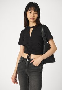 Svart crop-top med nøkkelhullskutt og samlet front. Kombinert med mørkegrå jeans og en svart skulderveske. Glatt stofftekstur.