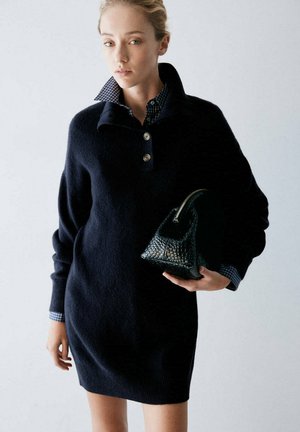 Une femme porte une robe-pull bleu marine sur une chemise à carreaux, tenant un petit sac à main noir texturé devant un fond clair uni.