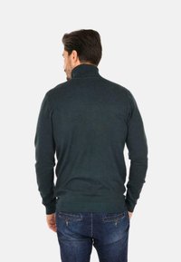 Maglione verde a maglia con colletto alto, texture a coste e design aderente, abbinato a jeans blu scuro. L'immagine mostra il retro dell'outfit.