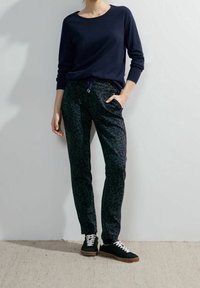 Camicia blu scuro a maniche lunghe con scollo rotondo, abbinata a pantaloni verde scuro con un motivo a macchie di leopardo, e sneakers nere.