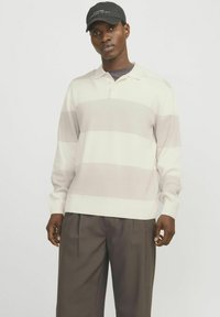 Strikpullover i creme med lysegrå vandrette striber, med en polo krave og lange ærmer, sammen med mørke bukser og en sort kasket.