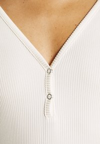 Primo piano di un indumento bianco a scollo a V, in tessuto a costine, con due bottoni metallici all'collo, che evidenzia la sua trama e i dettagli di design.