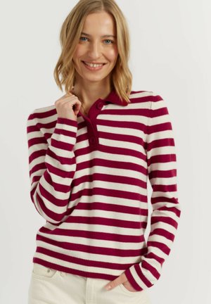 Vrouw die lacht, draagt een rood-wit gestreepte lange mouw polo met knopen en lichtgekleurde broek tegen een effen achtergrond.