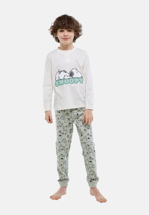 Enfant aux cheveux bouclés, debout pieds nus, portant un haut à manches longues blanc avec un motif Snoopy et un pantalon de pyjama clair à motif Snoopy.