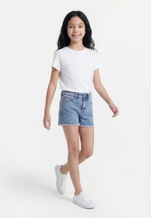 Pige gående, iført en hvid t-shirt, blå denimshorts med små farverige prikker og hvide sneakers mod en ensfarvet lys baggrund.