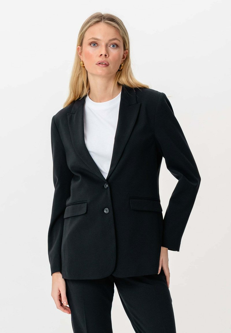 Femme blonde portant des boucles d'oreilles en or, un blazer noir ajusté à deux boutons, un pantalon noir et une chemise blanche sur fond blanc.