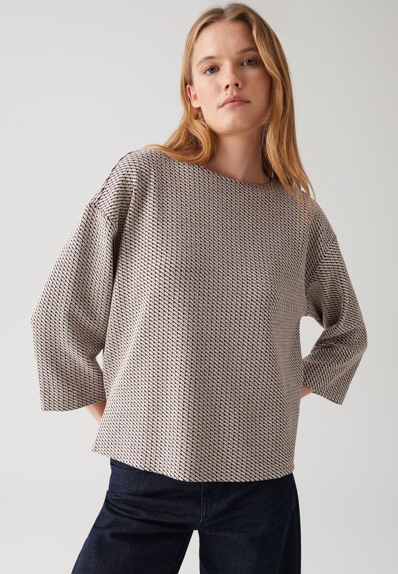 Femme jeune portant un pull ample beige et noir à motif avec des manches trois-quarts et un jean bleu foncé, se tenant les mains sur les hanches.