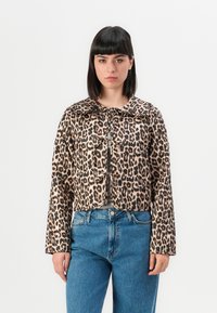 Kvinna med rakt mörkt hår som bär en kort leopardmönstrad jacka med knytdetaljer och högmidjade blå jeans, stående mot vit bakgrund.