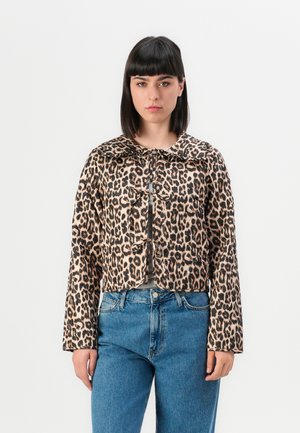 Vrouw met steil, donker haar, in een korte jasje met luipaardprint en striksluitingen, met hoge taille blauwe jeans, staand tegen een witte achtergrond.