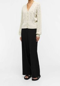 Cárdigan de punto color crema claro con un patrón de diamantes, con mangas largas, escote en V y cierre frontal de botones, combinado con pantalones de pierna ancha negros.