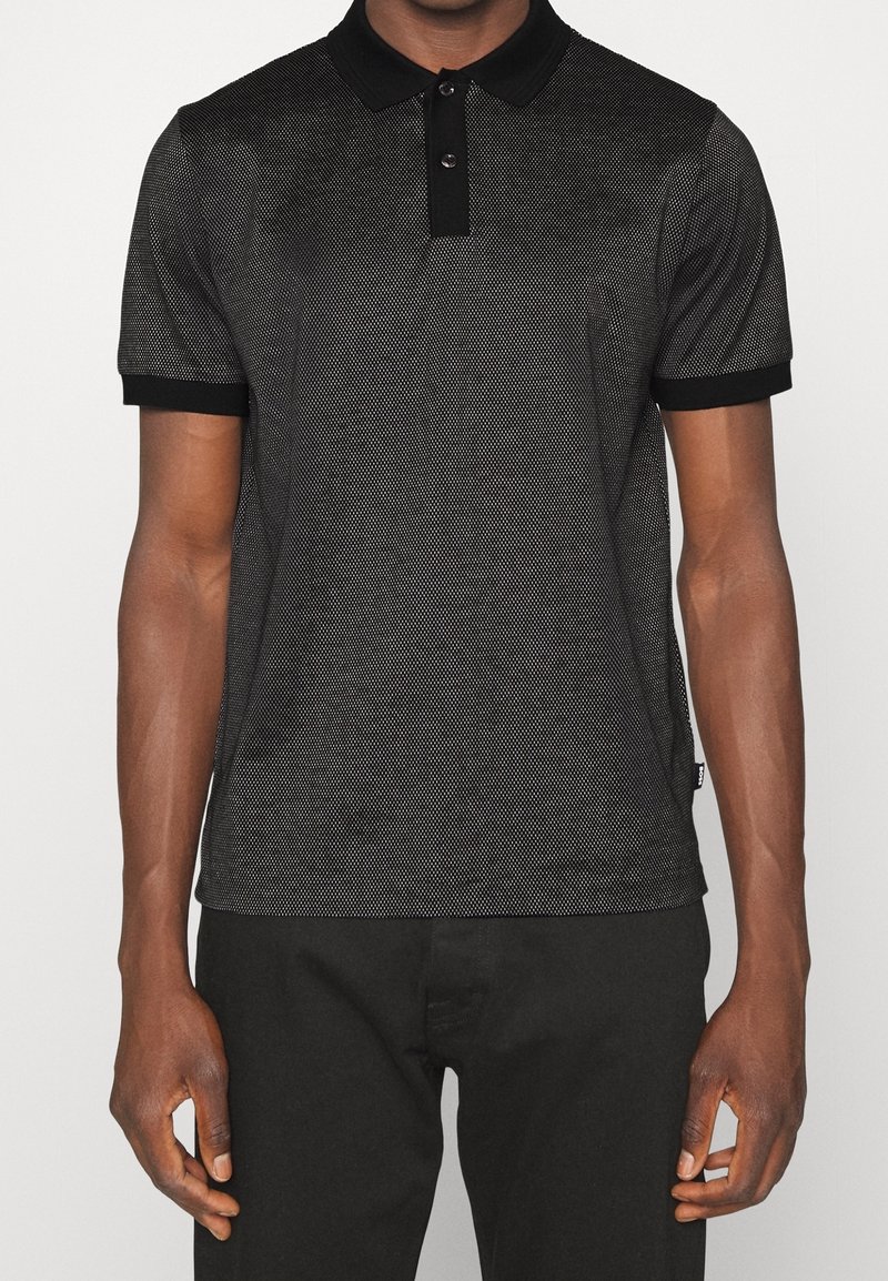 Homme portant un polo à manches courtes noir et gris à motifs, avec col et poignets noirs, associé à un pantalon noir.